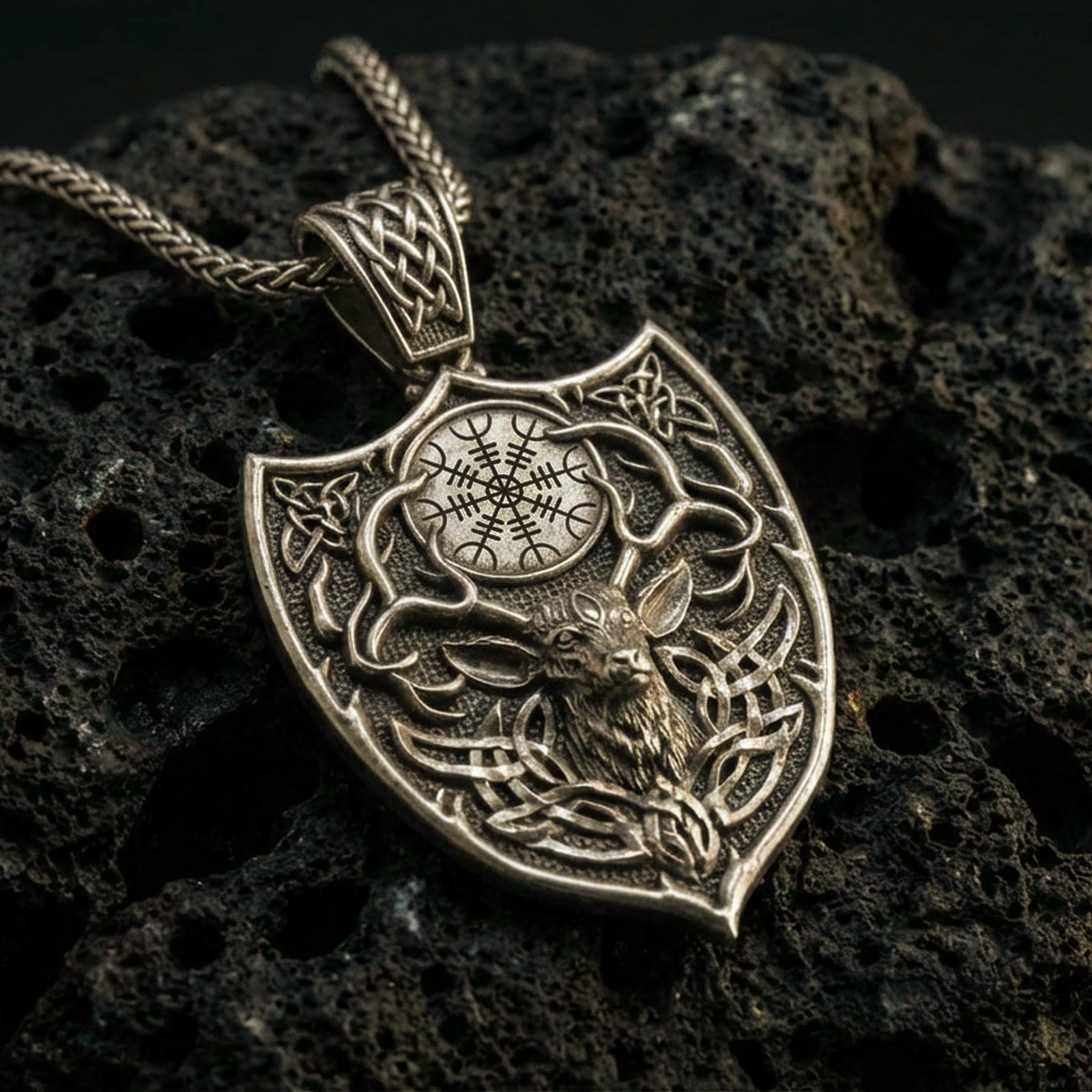 Bronze Celtic deer pendant with stag head motif, antler silhouette, and shield-shaped Norse design displayed on dark volcanic rock in a close-up luxury jewelry composition