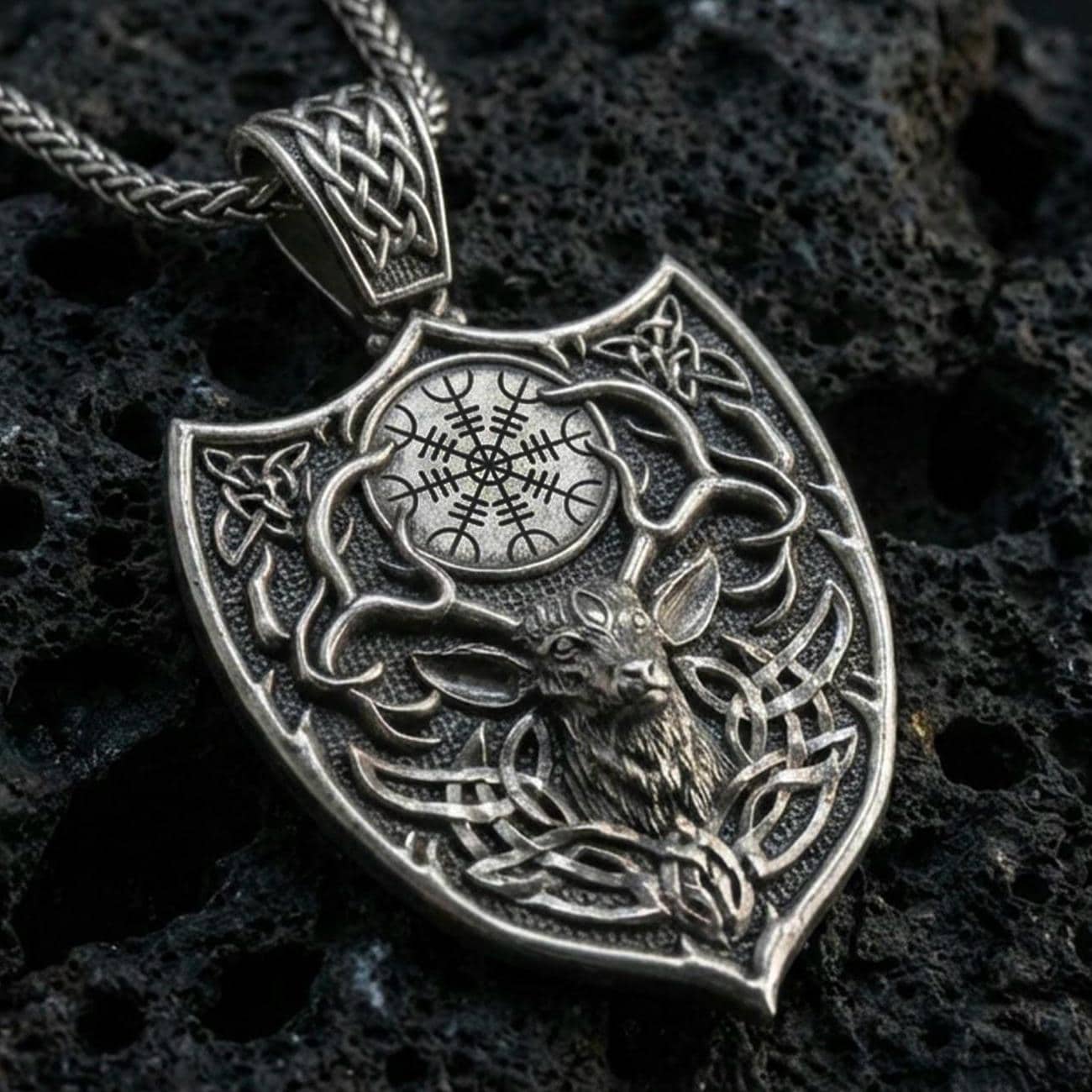 Oxidized Celtic deer pendant with stag head motif, antler silhouette, and shield-shaped Norse design displayed on dark volcanic rock in a close-up luxury jewelry composition