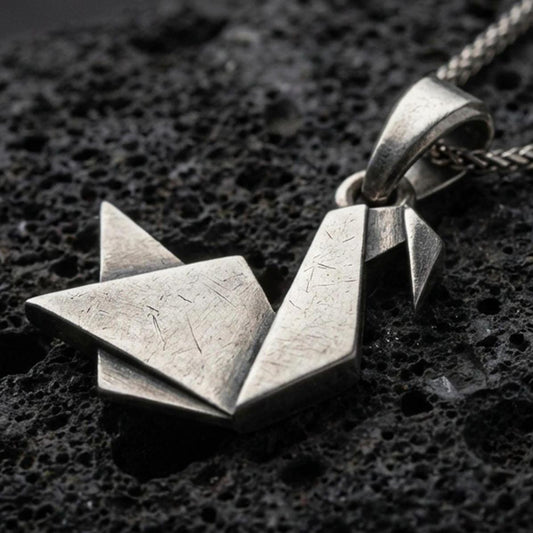 Oxidized origami swan pendant with folded geometric silhouette, minimalist angular lines, and elegant bird design displayed on dark volcanic rock in a close-up luxury jewelry composition