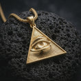 Load image into Gallery viewer, Gold plated all-seeing eye pendant necklace presented on porous volcanic rock, highlighting the symbolic triangle design in a luxurious close-up jewelry image.
