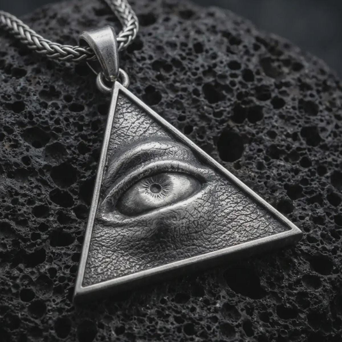 Sterling silver all-seeing eye pendant necklace with an oxidized finish resting on textured volcanic rock, captured in a close-up premium studio composition.