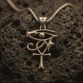 Load image into Gallery viewer, Bronze Eye of Ra and Ankh pendant necklace with an oxidized finish resting on textured volcanic rock, captured in a close-up premium studio composition.
