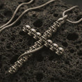 Load image into Gallery viewer, Bronze skull cross pendant necklace with an oxidized finish resting on textured volcanic rock, captured in a close-up premium studio composition.
