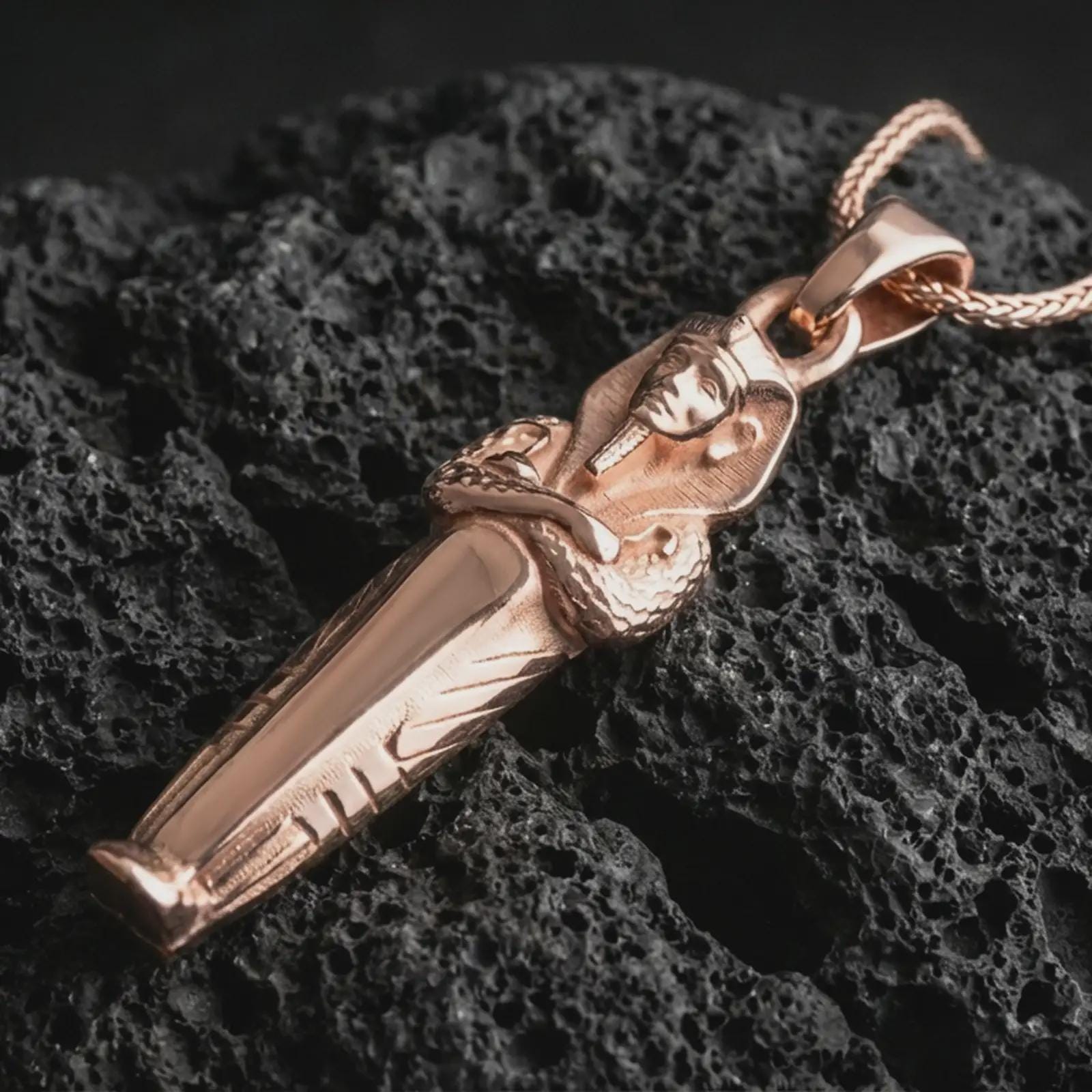 Rose gold plated King Tut pendant necklace on dark volcanic rock, photographed in a detailed premium studio macro view.