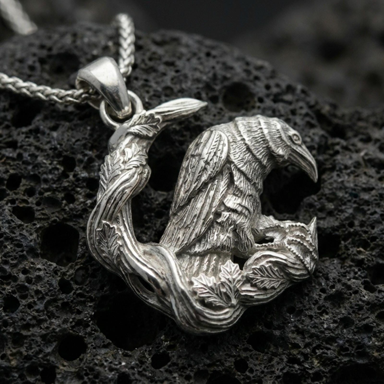 Polished silver Viking raven pendant necklace resting on black volcanic rock, captured in a refined macro product shot with intricate branch details.