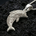 Load image into Gallery viewer, Polished silver dolphin pendant necklace displayed on black volcanic rock, shown in a refined macro product shot with engraved geometric detail.
