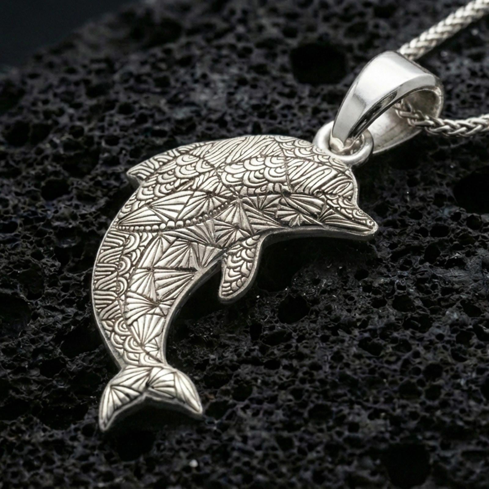 Polished silver dolphin pendant necklace displayed on black volcanic rock, shown in a refined macro product shot with engraved geometric detail.