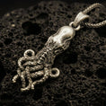 Load image into Gallery viewer, Bronze octopus pendant necklace with an oxidized finish resting on textured volcanic rock, captured in a close-up premium studio composition.
