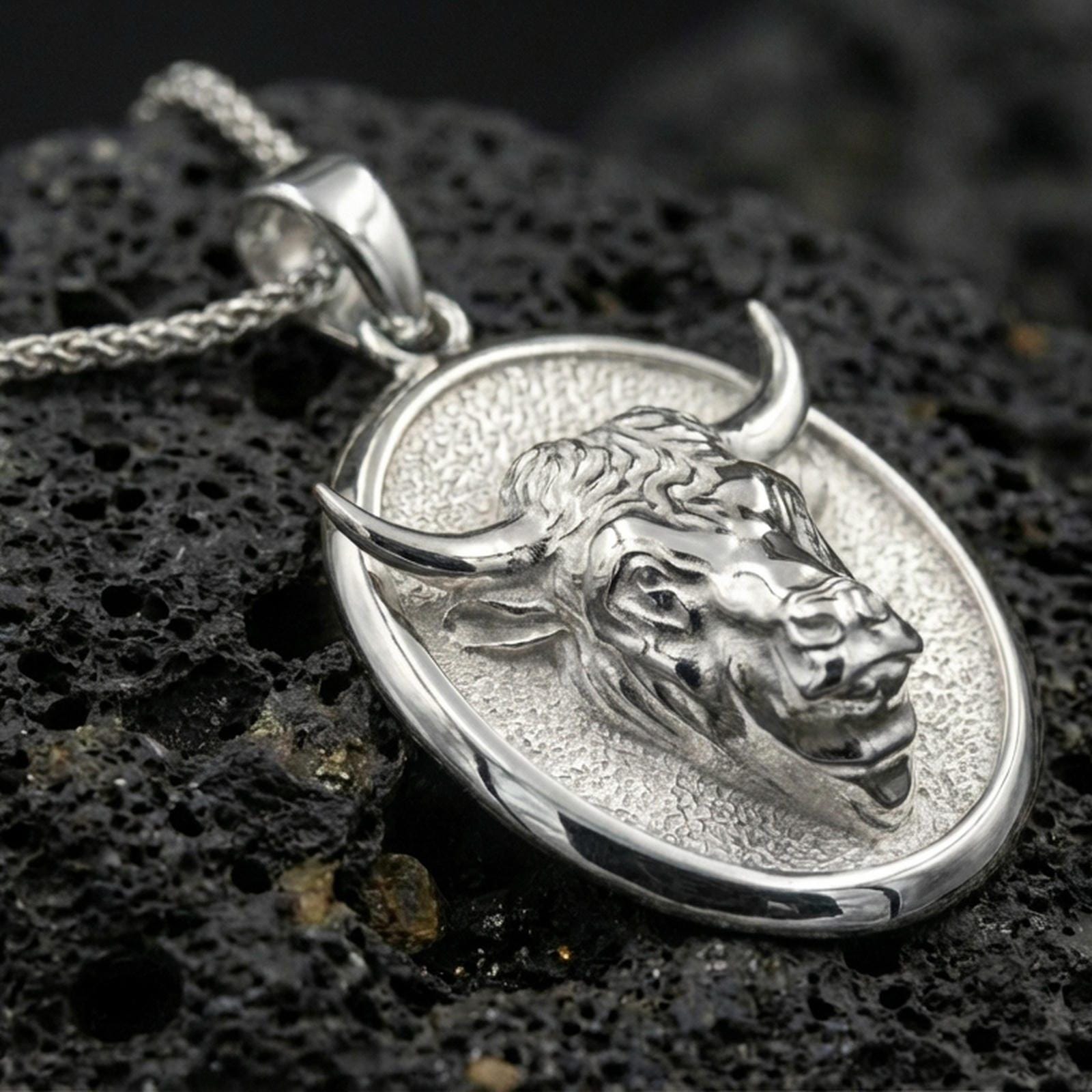 Polished silver bull pendant necklace displayed on black volcanic rock, shown in a refined macro product shot with sculpted horn and facial detail.