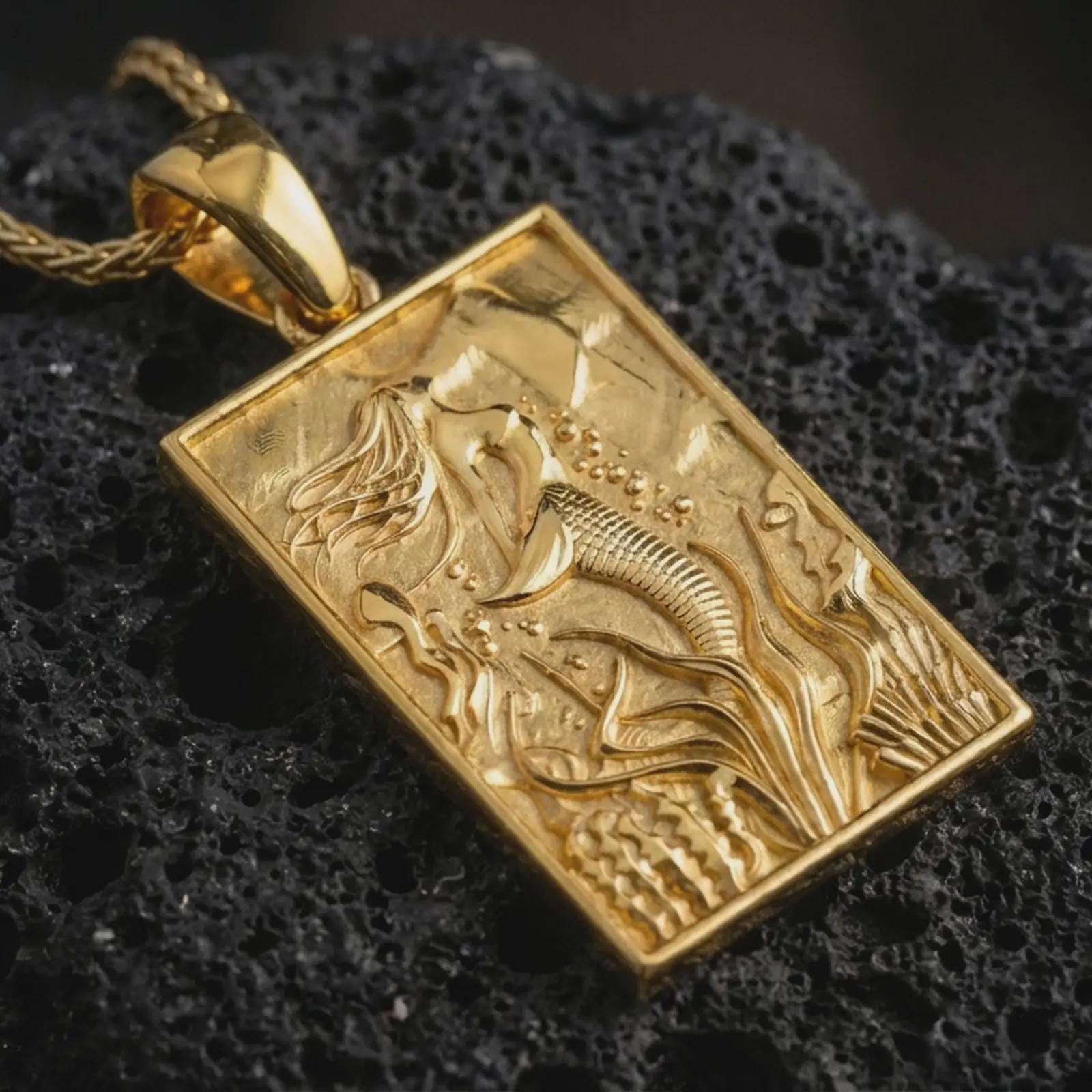 Gold plated mermaid pendant necklace presented on porous volcanic rock, highlighting the rectangular ocean-inspired design in a luxurious close-up jewelry image.
