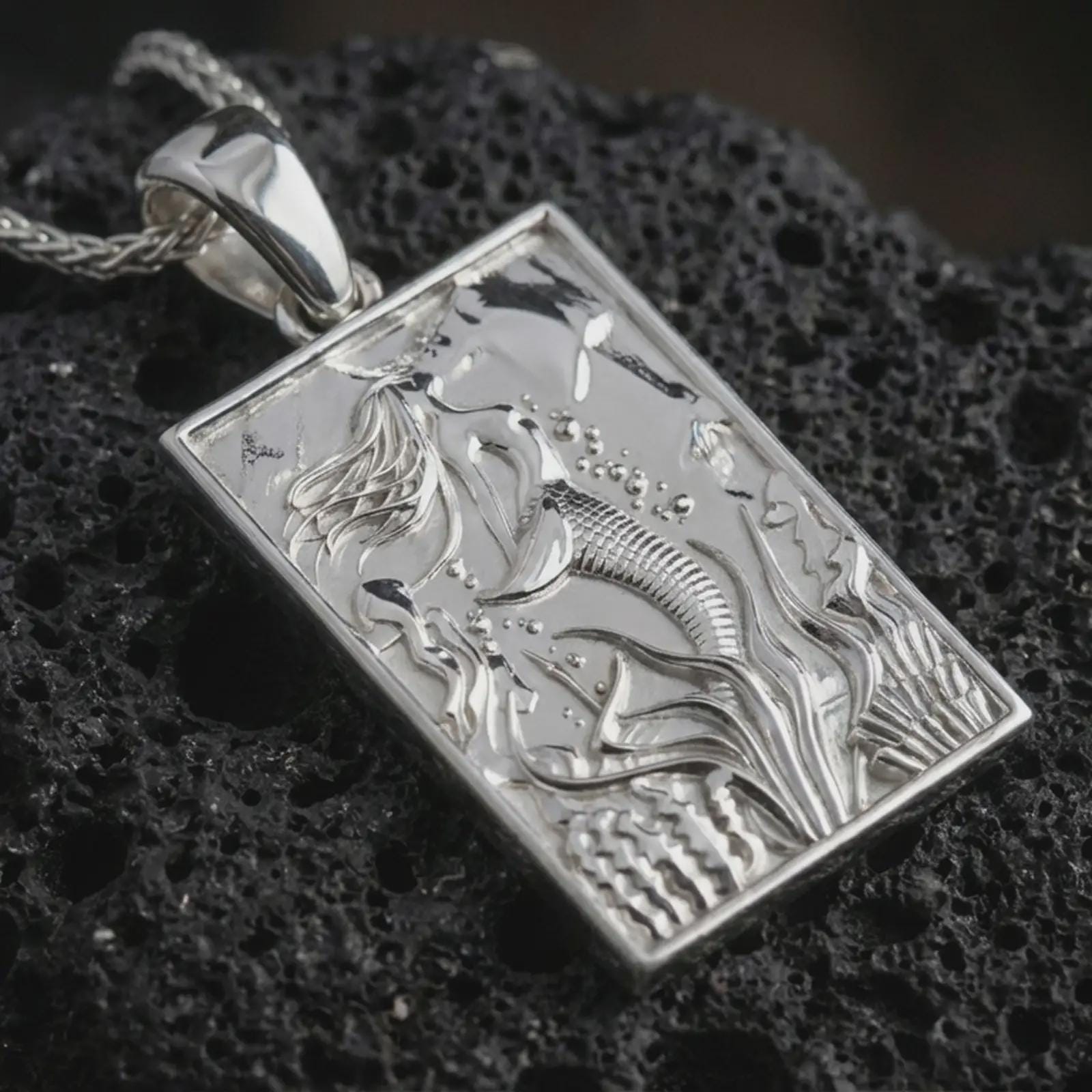 Polished silver mermaid pendant necklace displayed on black volcanic rock, shown in a refined macro product shot with detailed siren figure and flowing underwater relief.