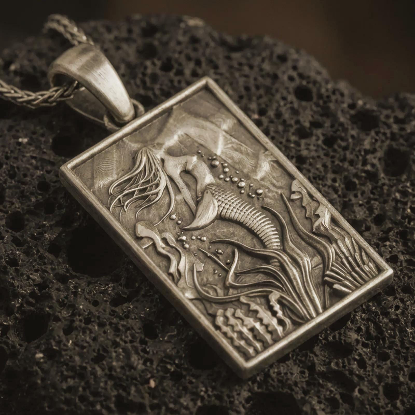 Bronze mermaid pendant necklace with an oxidized finish resting on textured volcanic rock, captured in a close-up premium studio composition.