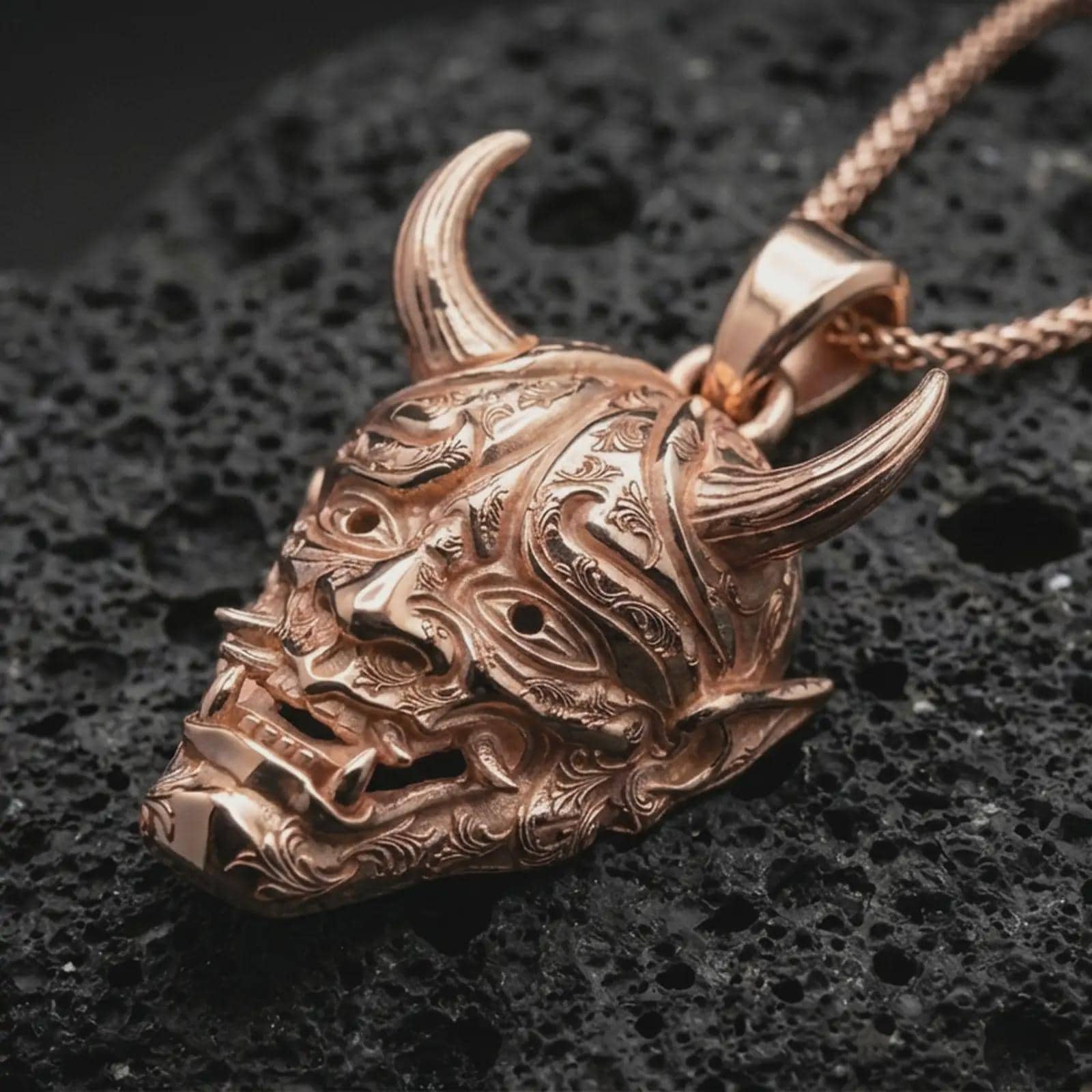 Rose gold plated Oni mask pendant necklace on dark volcanic rock, photographed in a detailed premium studio macro view.