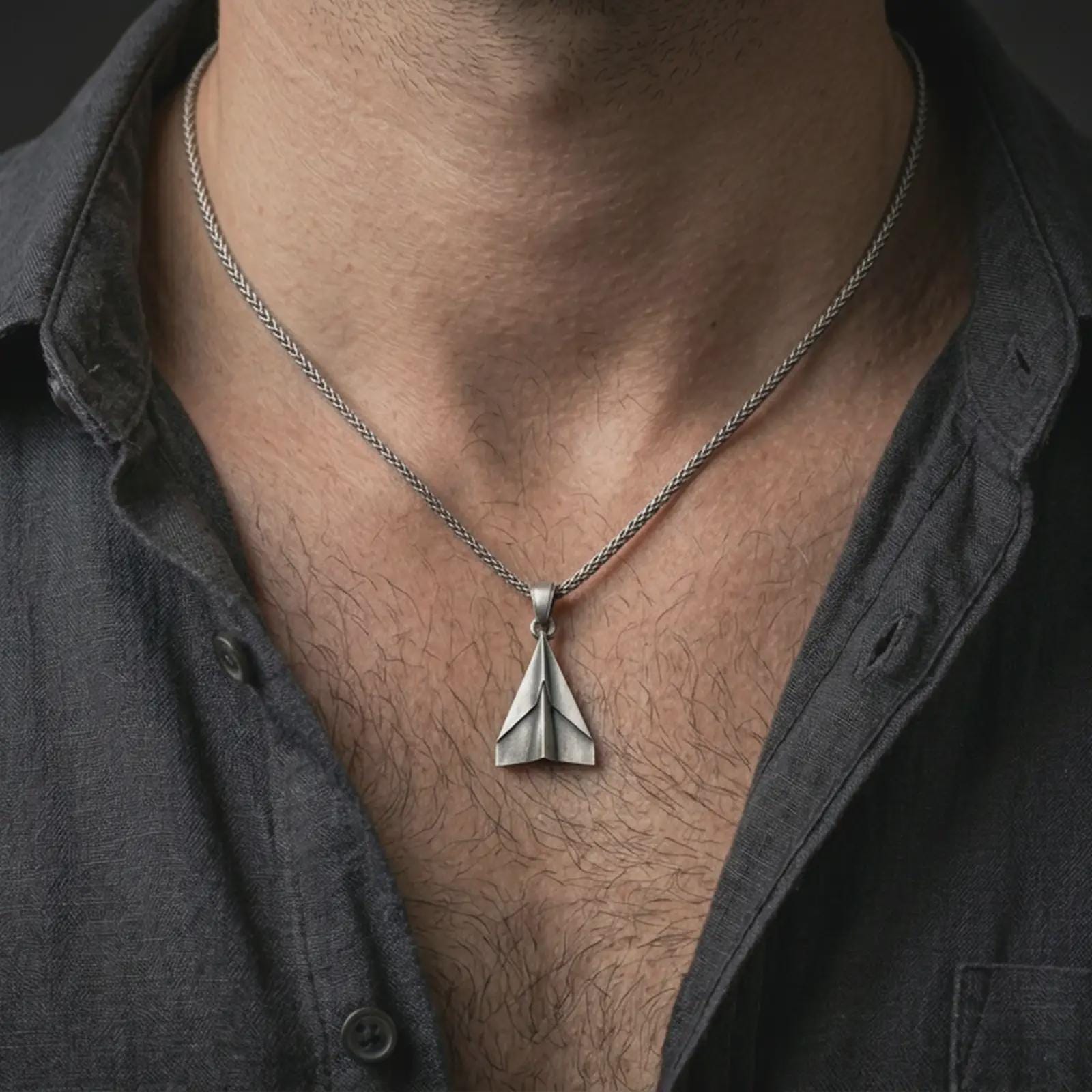 Oxidized origami airplane pendant necklace worn on the neck, styled in a dark lifestyle portrait with an open shirt and masculine presentation.