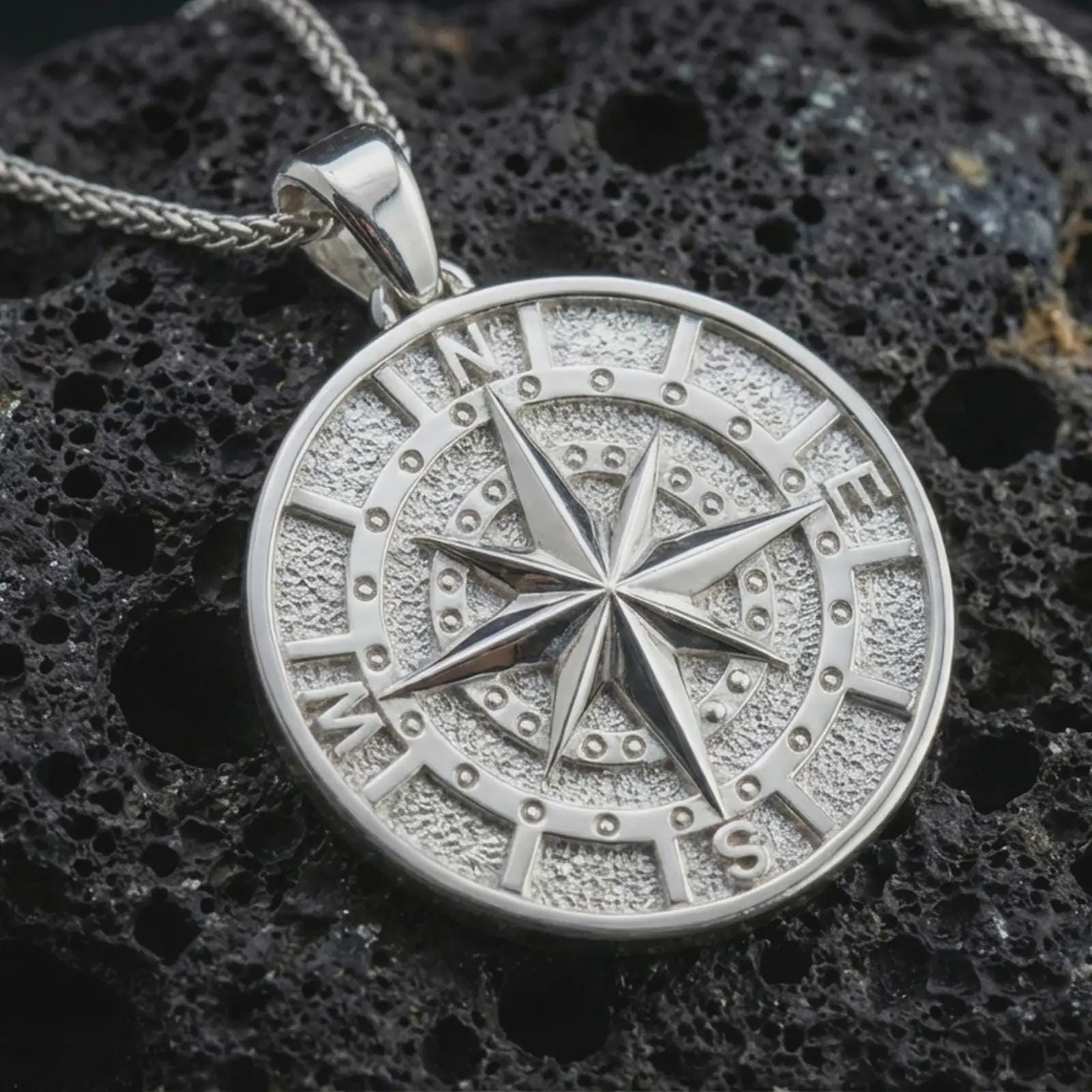 Polished silver compass pendant necklace displayed on black volcanic rock, shown in a refined macro product shot with detailed cardinal directions and raised star relief.