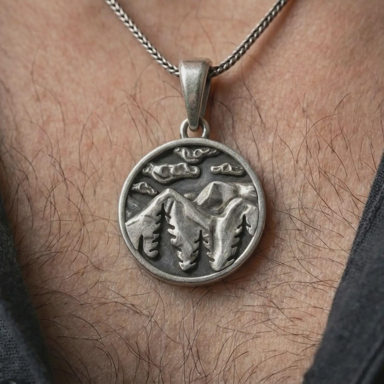 Oxidized mountain landscape pendant necklace worn on the neck, styled in a dark lifestyle portrait with an open shirt and masculine presentation.