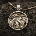 Load image into Gallery viewer, Bronze mountain landscape pendant necklace with an oxidized finish resting on textured volcanic rock, captured in a close-up premium studio composition.
