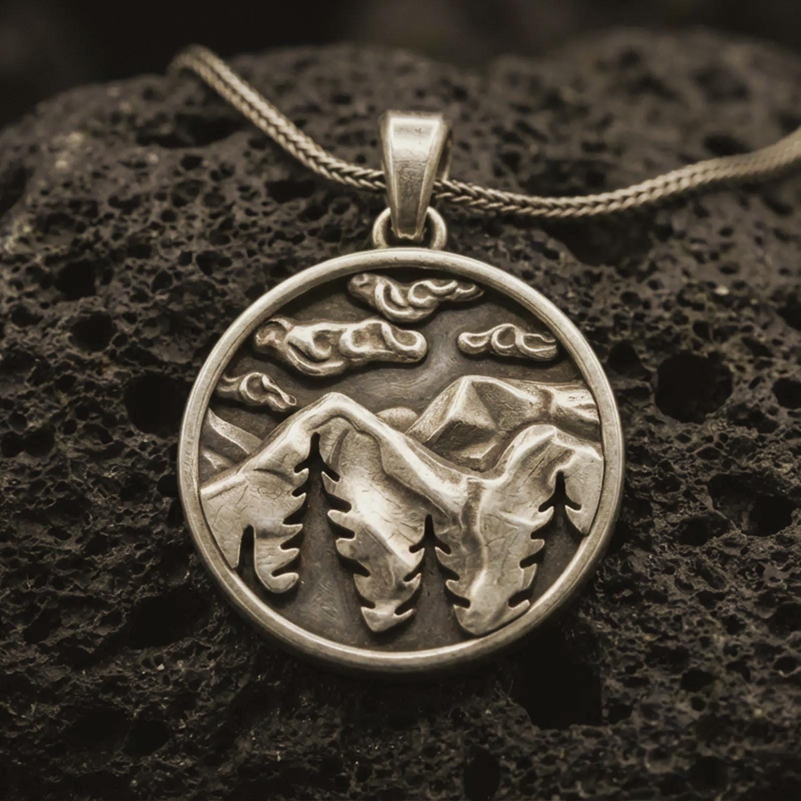 Bronze mountain landscape pendant necklace with an oxidized finish resting on textured volcanic rock, captured in a close-up premium studio composition.