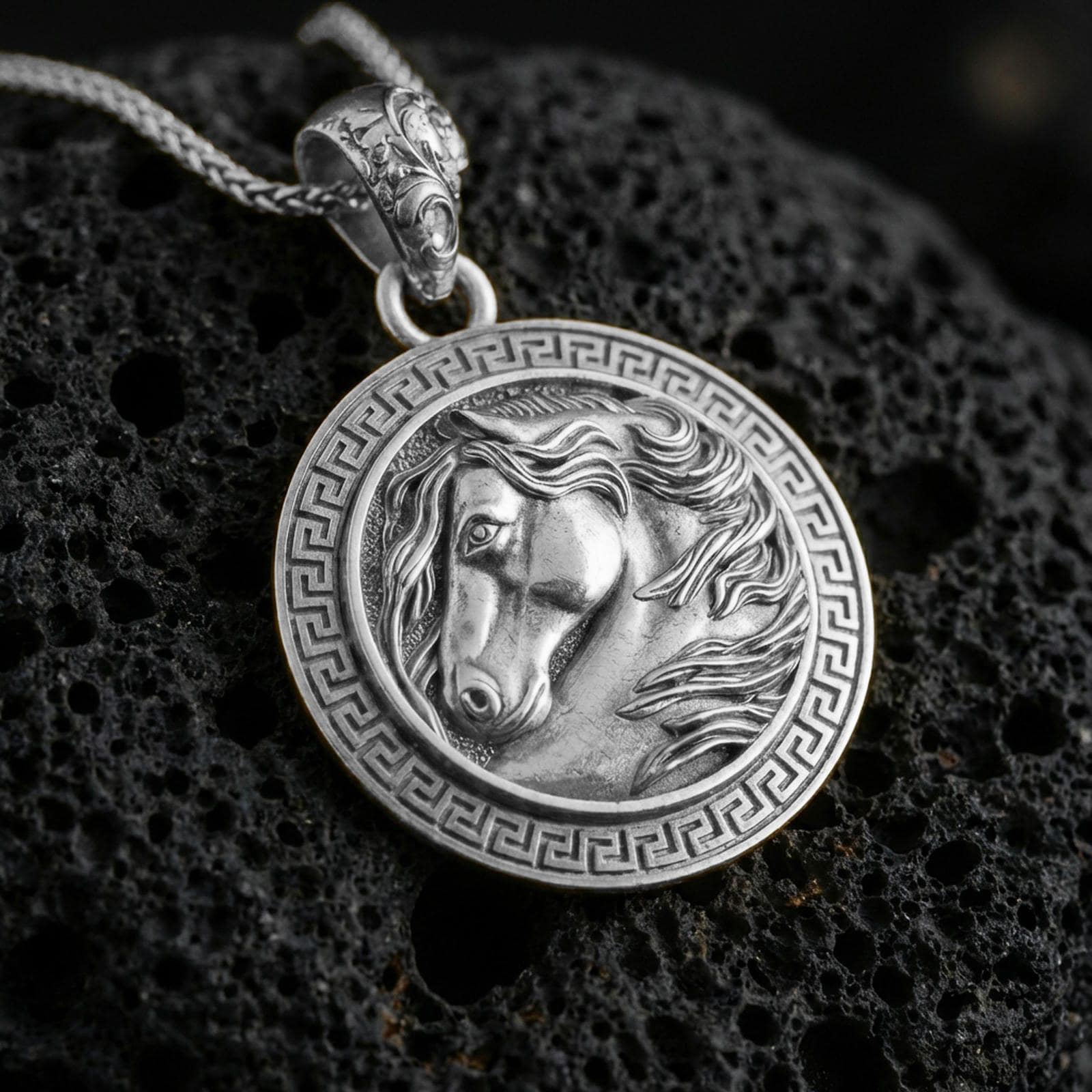 Polished silver horse head pendant necklace displayed on black volcanic rock, shown in a refined macro product shot with Greek key border detail.