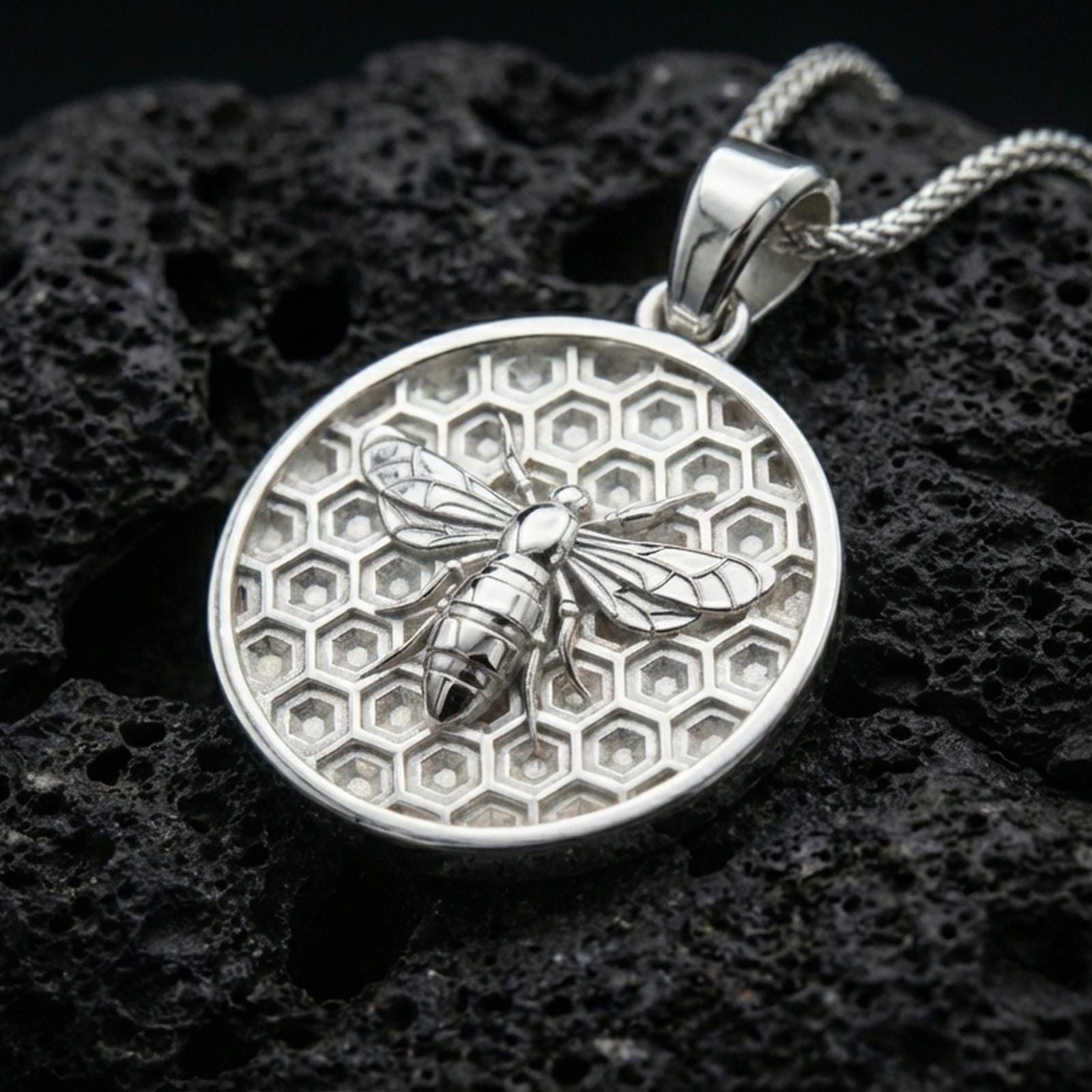 Polished silver bee pendant necklace displayed on black volcanic rock, shown in a refined macro product shot with honeycomb background detail.