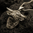 Load image into Gallery viewer, Bronze buffalo head pendant necklace with an oxidized finish resting on textured volcanic rock, captured in a close-up premium studio composition.

