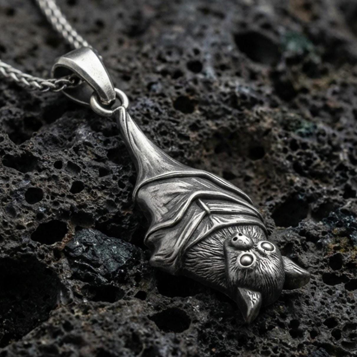 Sterling silver hanging bat pendant necklace with an oxidized finish resting on textured volcanic rock, captured in a close-up premium studio composition.