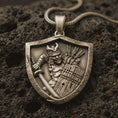 Load image into Gallery viewer, Bronze samurai shield pendant necklace with an oxidized finish resting on textured volcanic rock, captured in a close-up premium studio composition.
