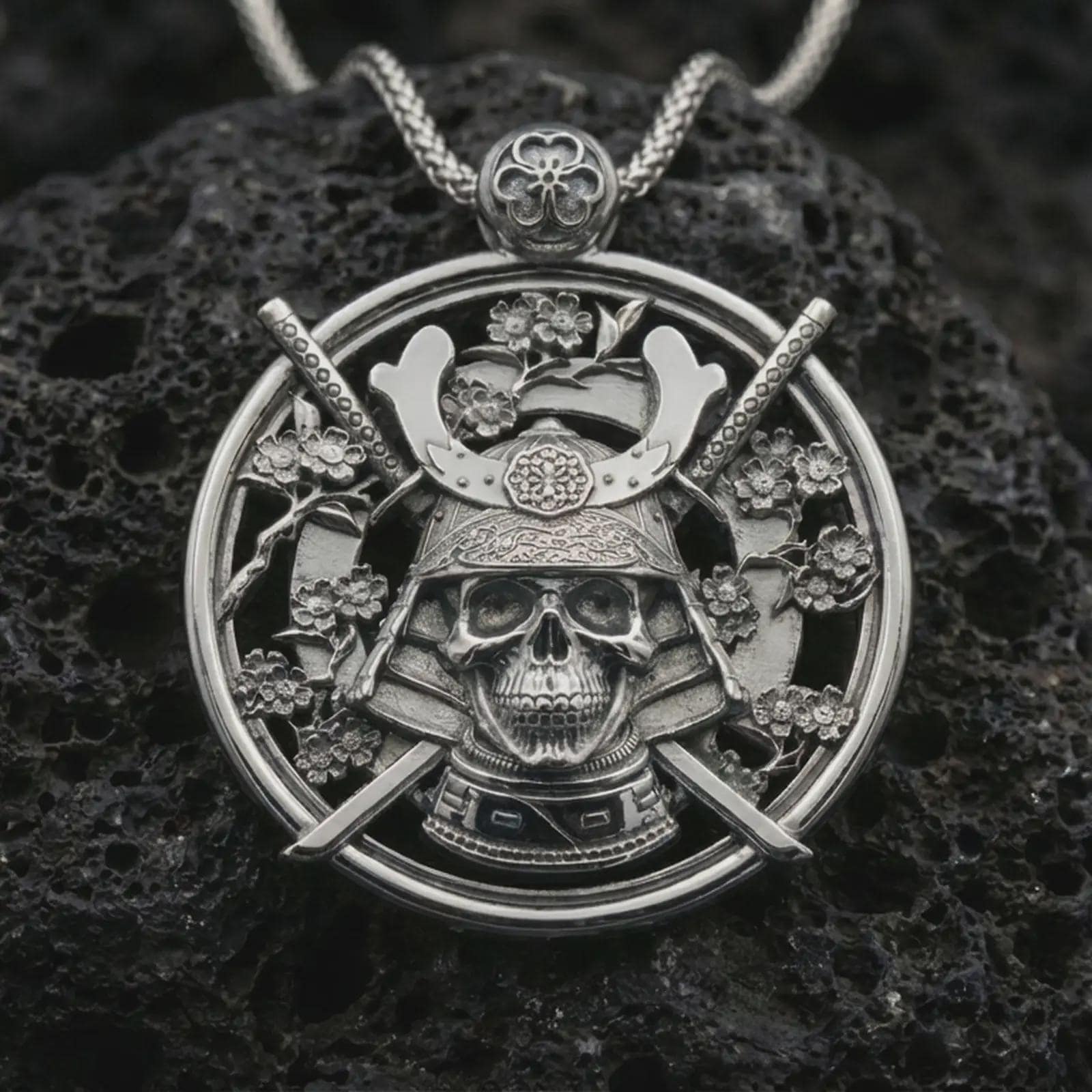 Polished silver samurai skull pendant necklace displayed on black volcanic rock, shown in a refined macro product shot with katana swords, floral accents, and armored helmet detail.