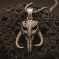 Load image into Gallery viewer, Bronze Mythosaur skull pendant necklace with an oxidized finish resting on textured volcanic rock, captured in a close-up premium studio composition.
