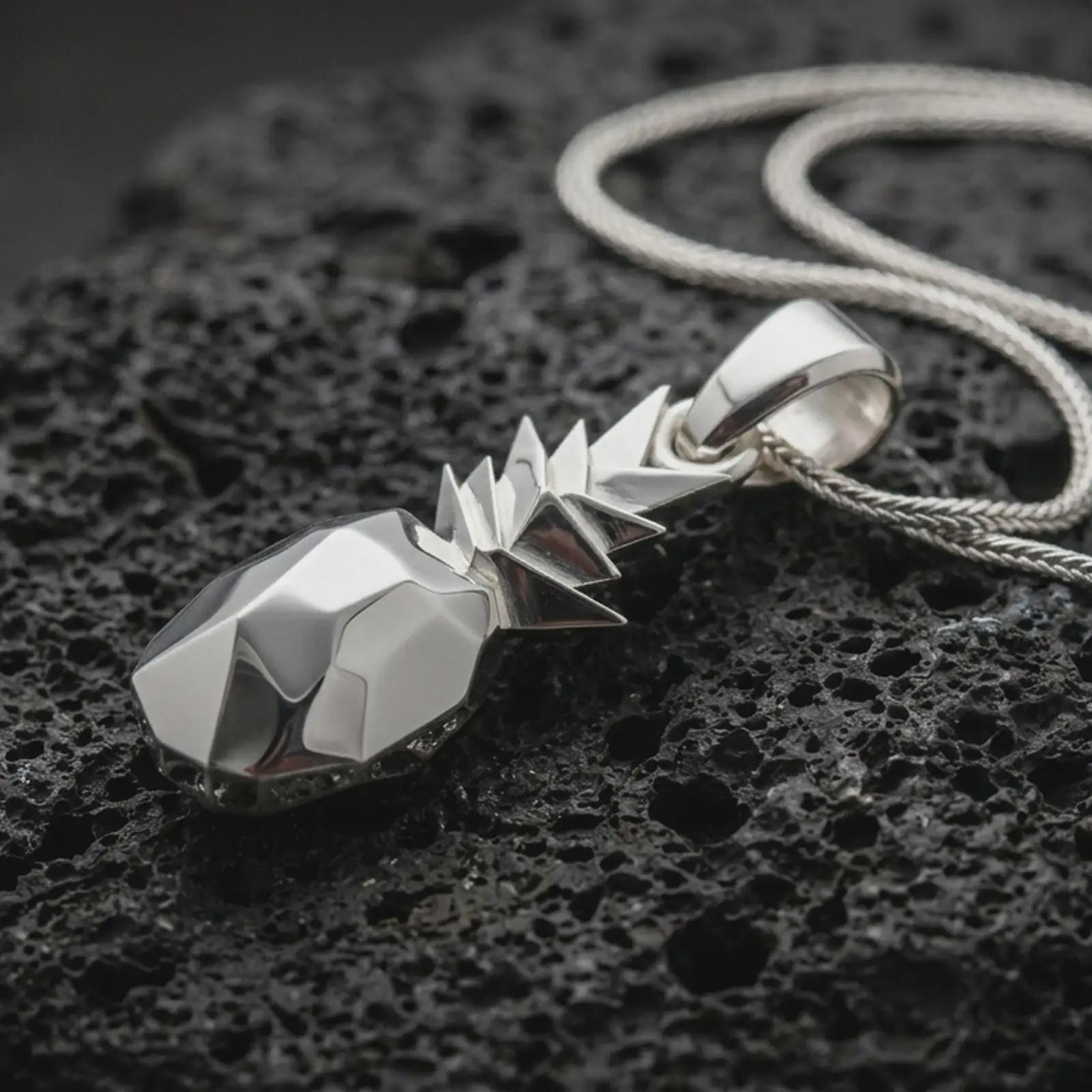 Polished silver origami pineapple pendant necklace displayed on black volcanic rock, shown in a refined macro product shot with faceted geometric body and sharp folded leaf crown detail.