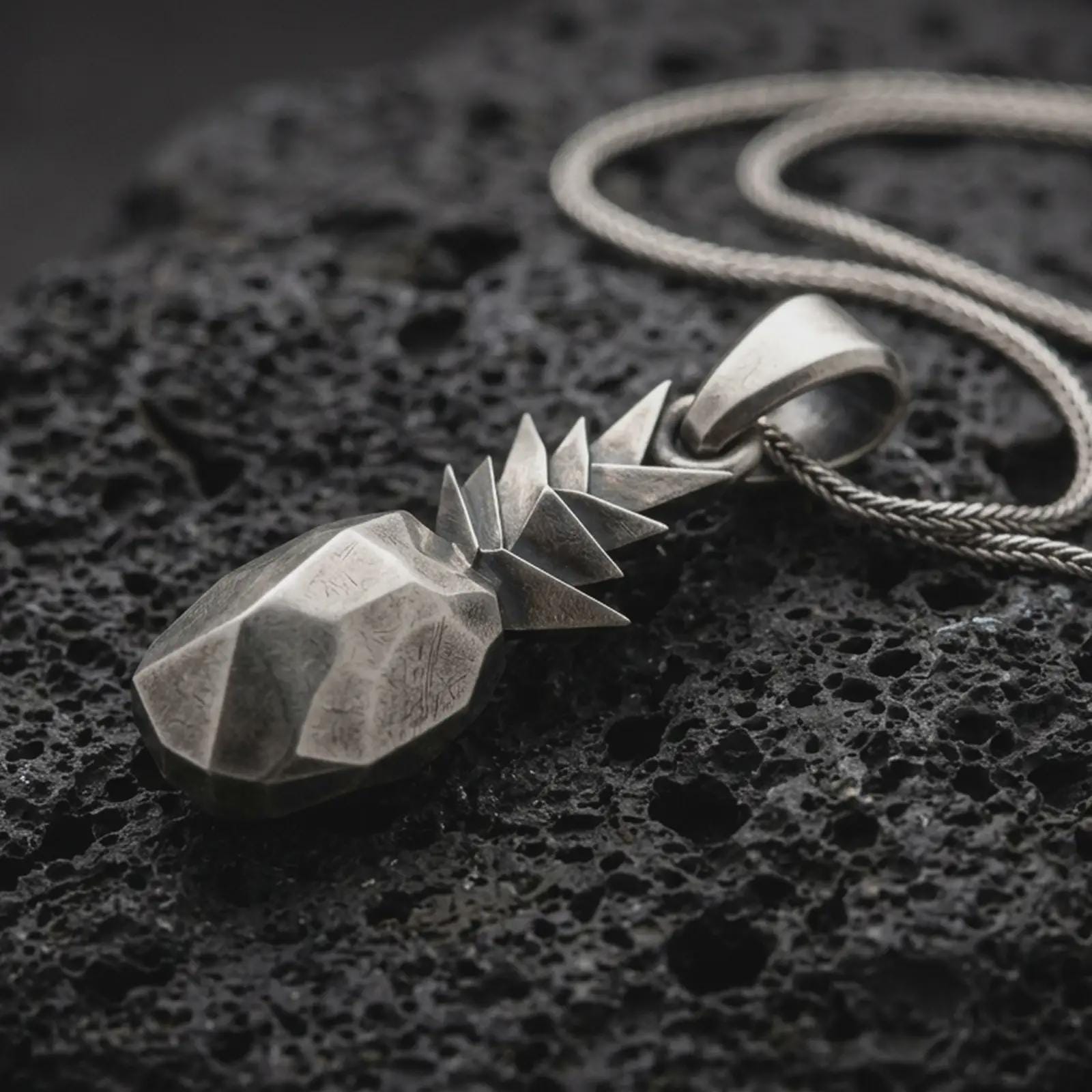 Sterling silver origami pineapple pendant necklace with an oxidized finish resting on textured volcanic rock, captured in a close-up premium studio composition.