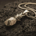 Load image into Gallery viewer, Bronze origami pineapple pendant necklace with an oxidized finish resting on textured volcanic rock, captured in a close-up premium studio composition.

