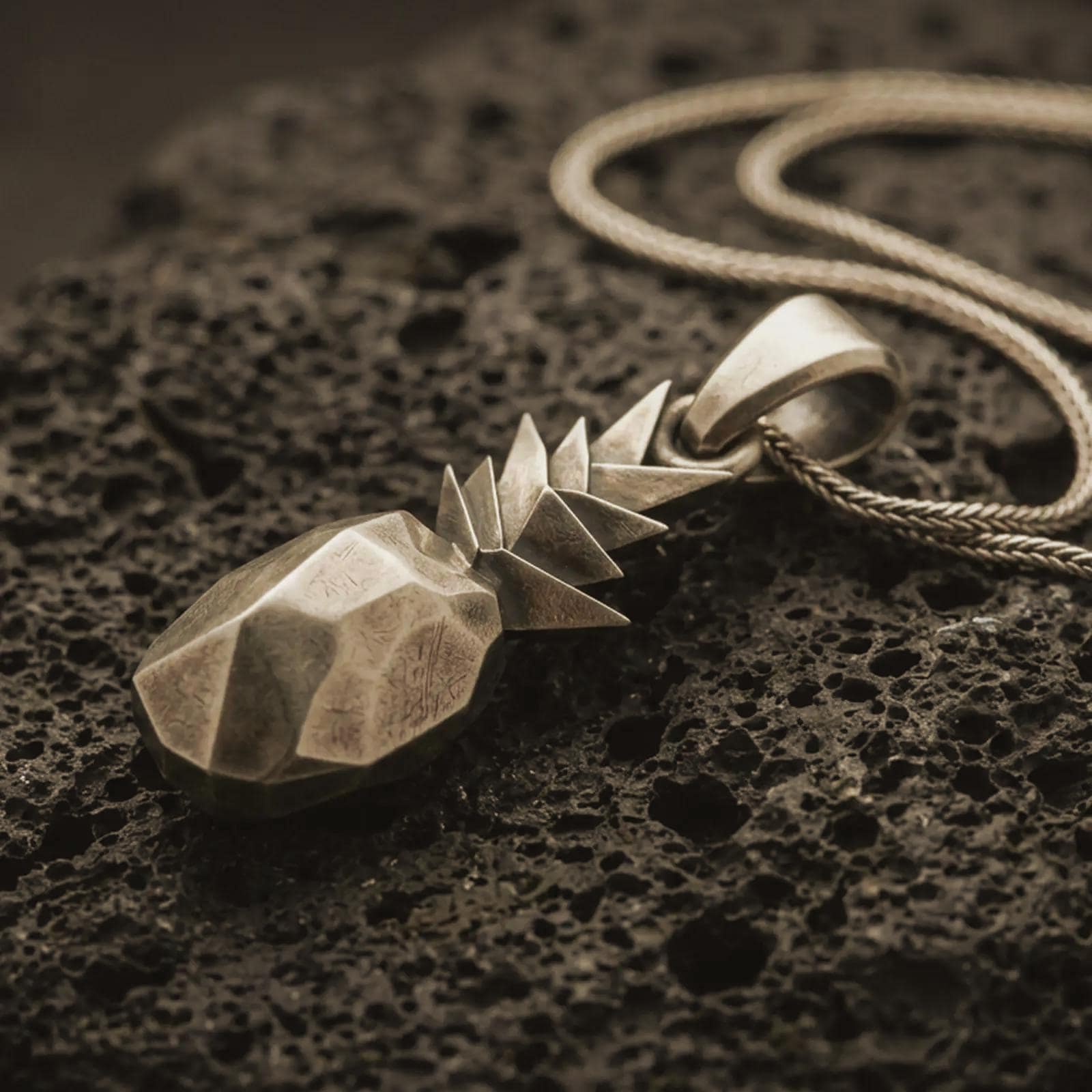 Bronze origami pineapple pendant necklace with an oxidized finish resting on textured volcanic rock, captured in a close-up premium studio composition.