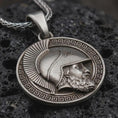 Load image into Gallery viewer, Sterling silver Spartan helmet pendant necklace with an oxidized finish resting on textured volcanic rock, captured in a close-up premium studio composition.
