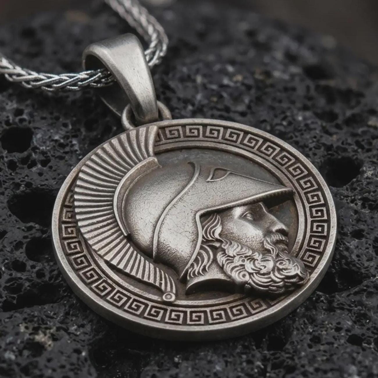Sterling silver Spartan helmet pendant necklace with an oxidized finish resting on textured volcanic rock, captured in a close-up premium studio composition.