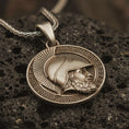 Load image into Gallery viewer, Bronze Spartan helmet pendant necklace with an oxidized finish resting on textured volcanic rock, captured in a close-up premium studio composition.
