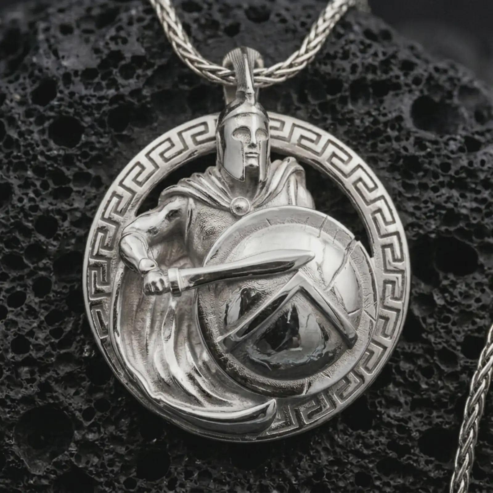 Polished silver Spartan warrior pendant necklace displayed on black volcanic rock, shown in a refined macro product shot with helmeted soldier, sword, shield, and Greek key border detail.
