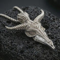 Load image into Gallery viewer, Polished silver Baphomet skull pendant necklace displayed on black volcanic rock, shown in a refined macro product shot with horned goat skull, engraved texture, and occult eye detail.
