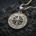 Load image into Gallery viewer, Polished silver compass rose pendant necklace displayed on black volcanic rock, shown in a refined macro product shot with cardinal direction letters and sharp navigational star detail.
