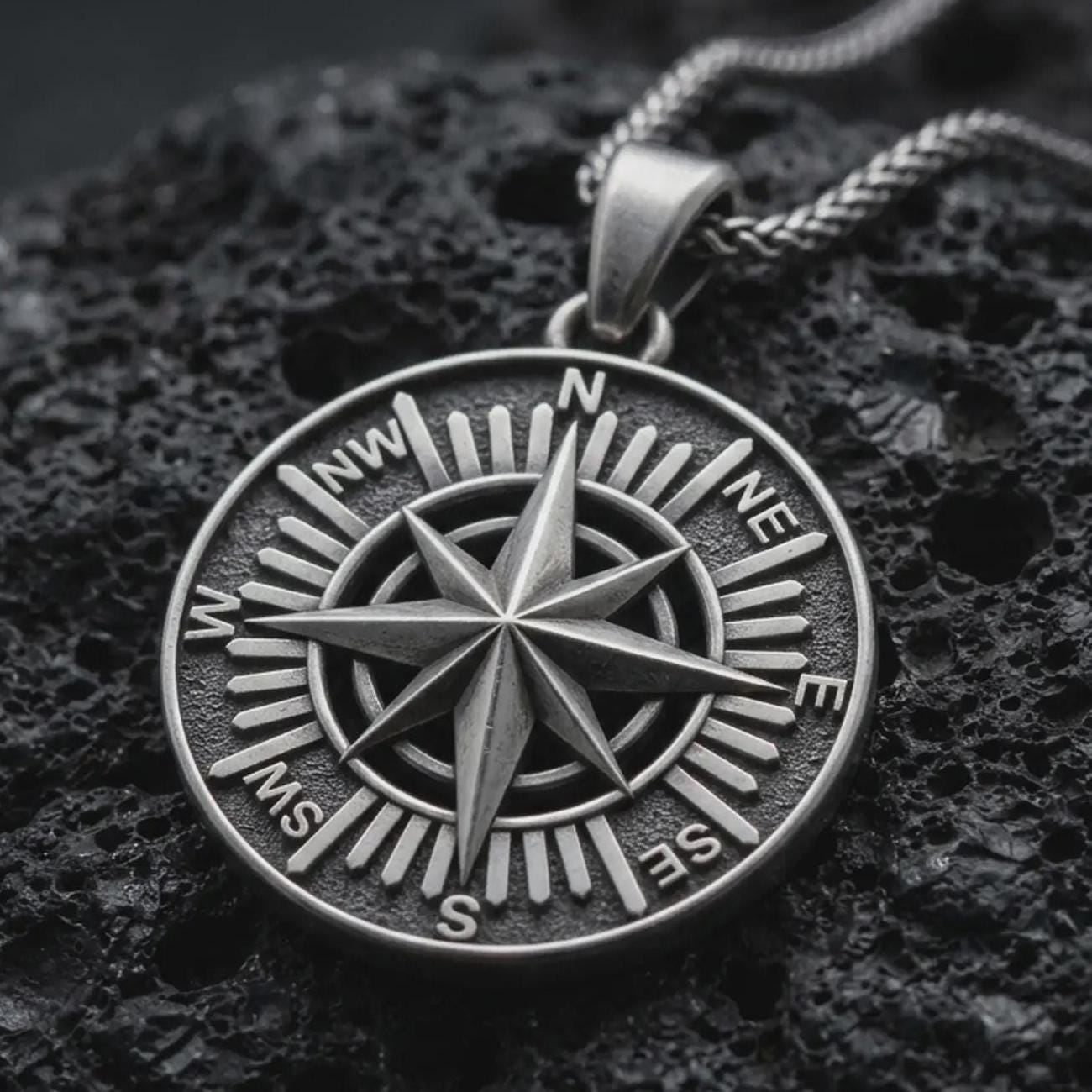 Sterling silver compass rose pendant necklace with an oxidized finish resting on textured volcanic rock, captured in a close-up premium studio composition.