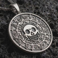 Load image into Gallery viewer, Sterling silver cursed Aztec coin pendant necklace with an oxidized finish resting on dark volcanic stone, captured in a close-up studio image with skull medallion and engraved tribal border details.
