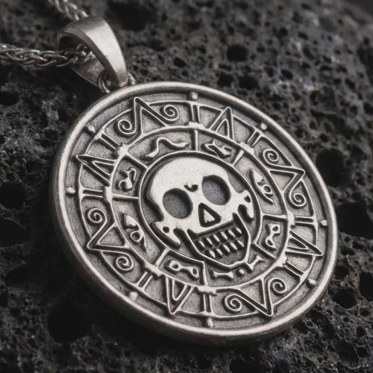 Sterling silver cursed Aztec coin pendant necklace with an oxidized finish resting on dark volcanic stone, captured in a close-up studio image with skull medallion and engraved tribal border details.