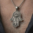 Load image into Gallery viewer, Oxidized Hamsa hand pendant necklace worn on the neck, styled in a dark lifestyle portrait with an open shirt and elegant presentation.
