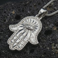 Load image into Gallery viewer, Polished silver Hamsa hand pendant necklace displayed on black volcanic rock, shown in a refined macro product shot with protective evil eye detail and beaded border relief.
