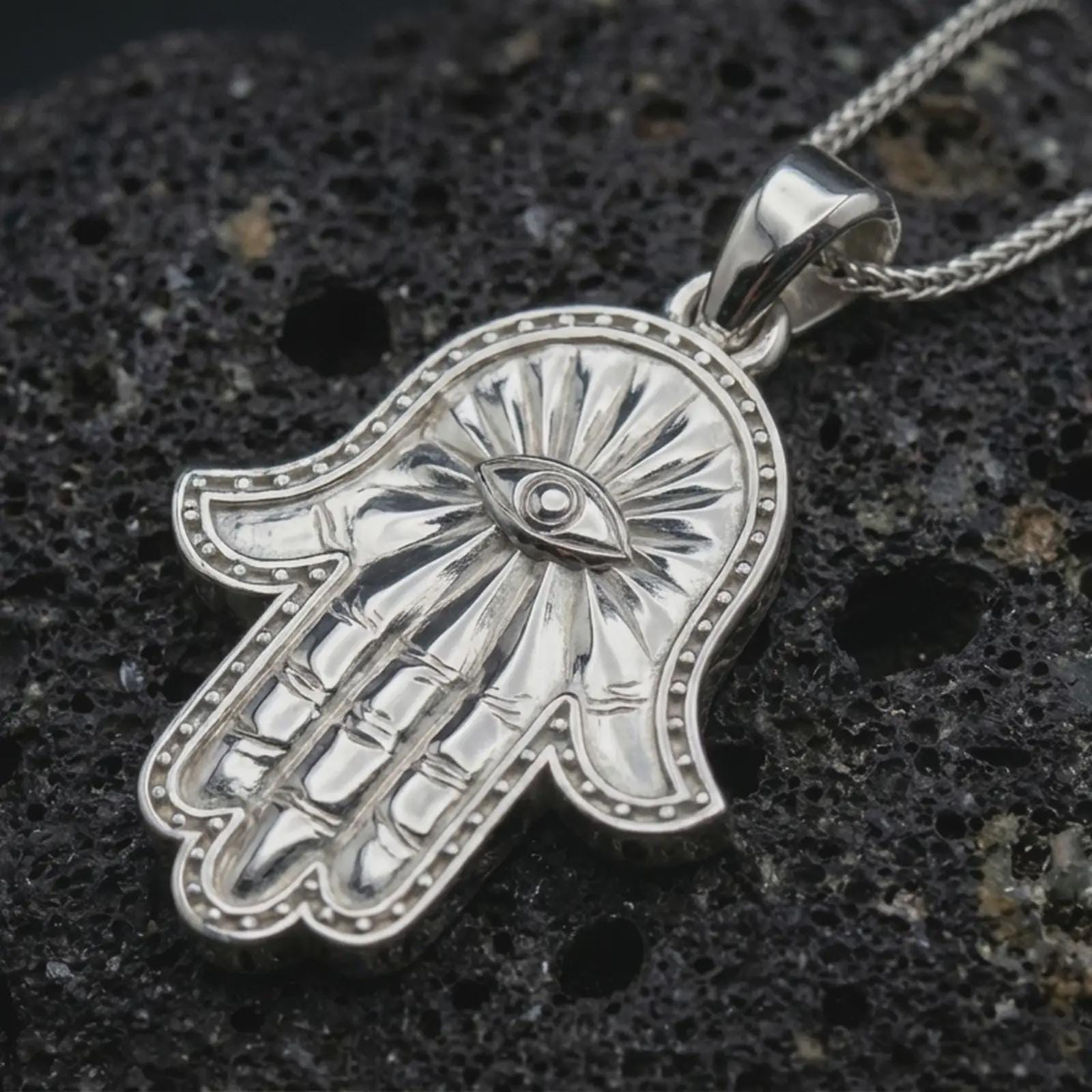 Polished silver Hamsa hand pendant necklace displayed on black volcanic rock, shown in a refined macro product shot with protective evil eye detail and beaded border relief.