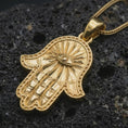 Load image into Gallery viewer, Gold plated Hamsa hand pendant necklace presented on porous volcanic rock, highlighting the symbolic palm design in a luxurious close-up jewelry image.
