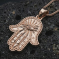 Load image into Gallery viewer, Rose gold plated Hamsa hand pendant necklace on dark volcanic rock, photographed in a detailed premium studio macro view.
