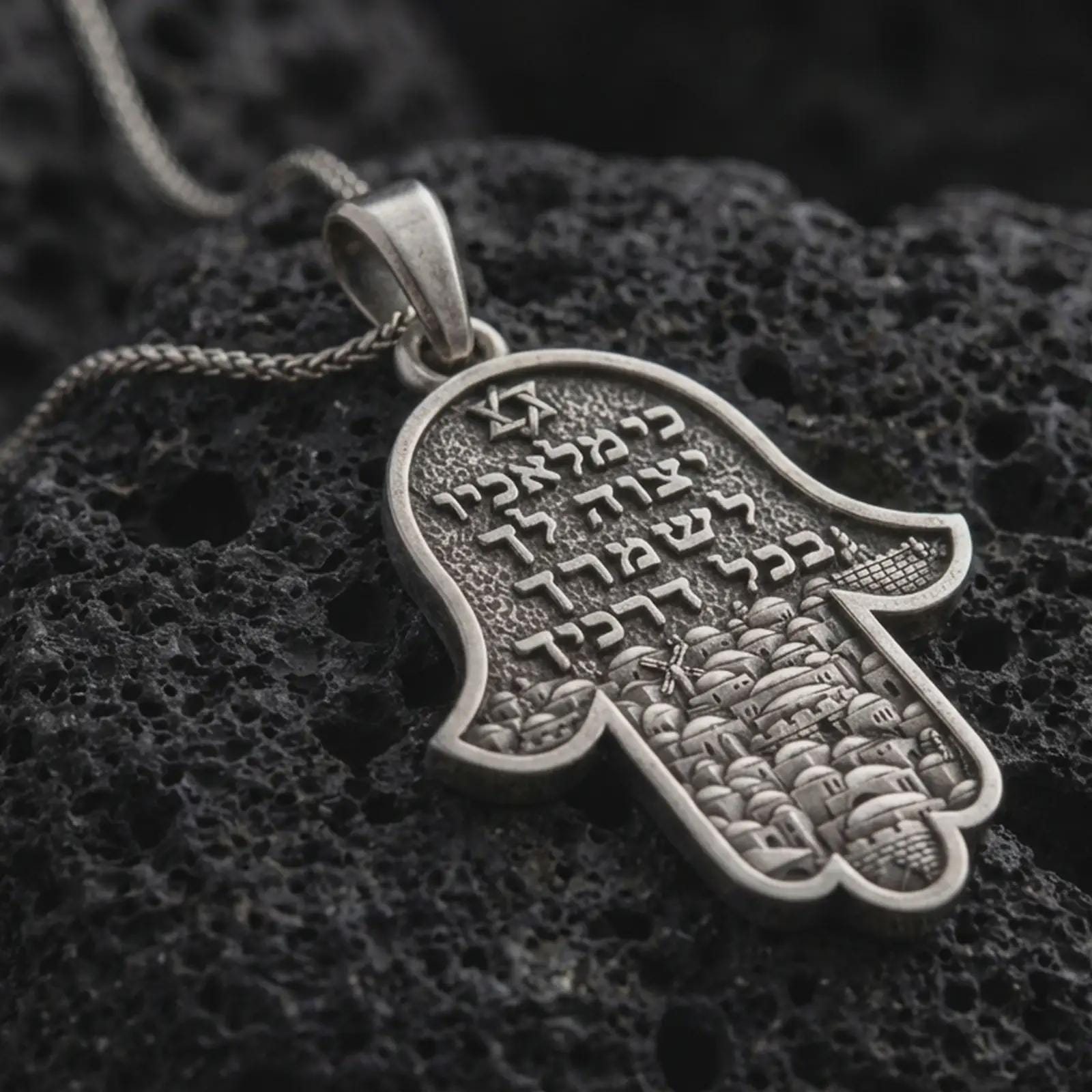 Sterling silver Jerusalem Hamsa hand pendant necklace with an oxidized finish resting on textured volcanic rock, captured in a close-up premium studio composition.