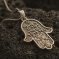 Load image into Gallery viewer, Bronze Jerusalem Hamsa hand pendant necklace with an oxidized finish resting on textured volcanic rock, captured in a close-up premium studio composition.
