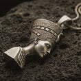 Load image into Gallery viewer, Bronze Nefertiti pendant necklace with an oxidized finish resting on textured volcanic rock, captured in a close-up premium studio composition.
